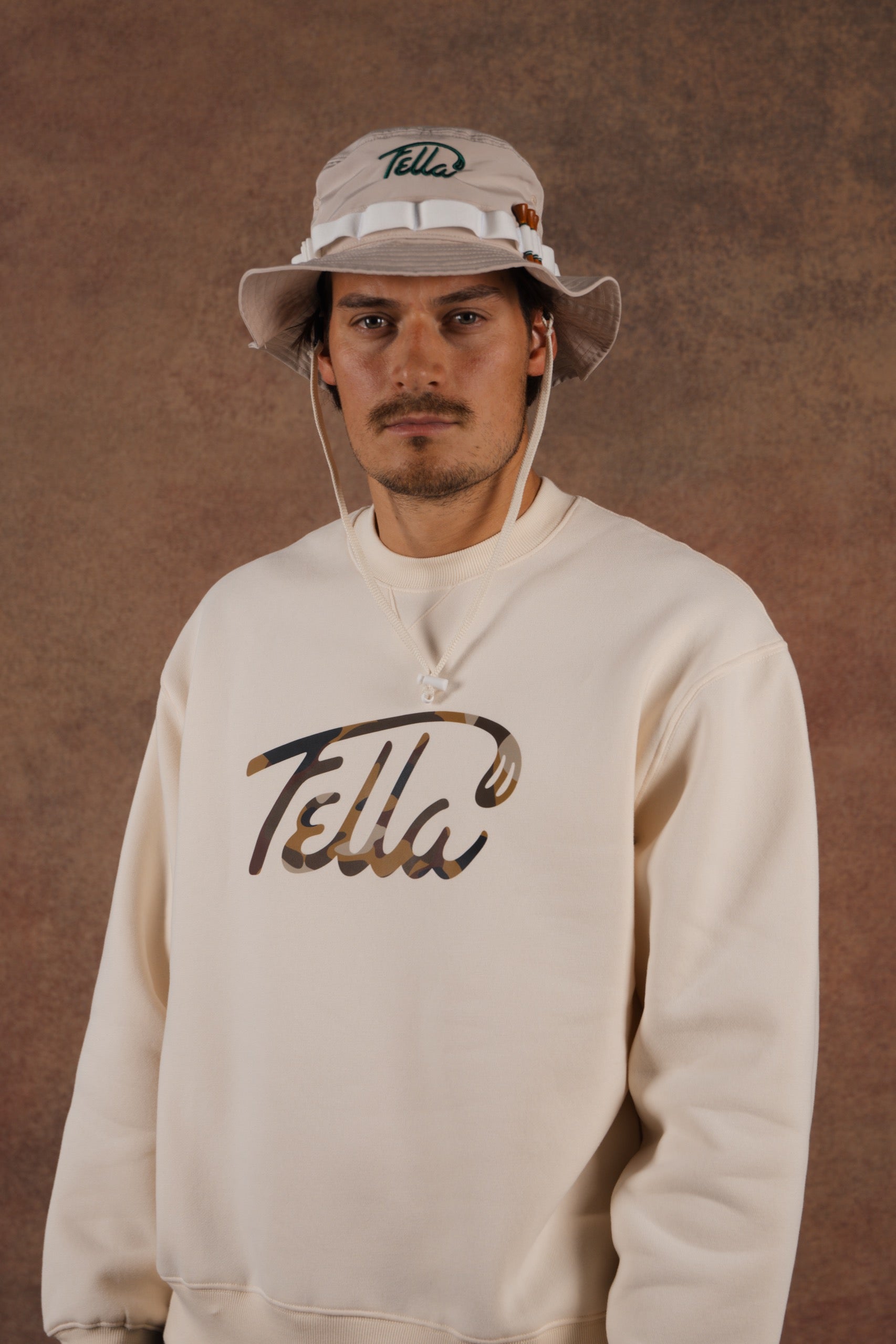 Man wearing a beige 'Fella' sweatshirt and cap against a brown background